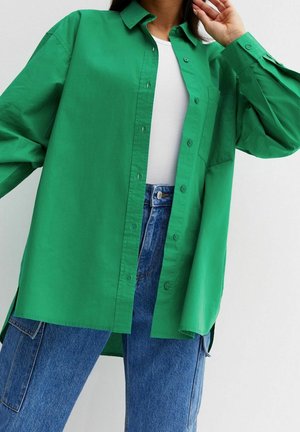 Personne portant une chemise boutonnée oversize vert vif par-dessus un haut côtelé blanc et un jean cargo en denim bleu taille haute, posant devant un fond blanc.