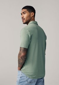 Mand med kort hår og ærme-tatovering iført grøn poloshirt og lyseblå jeans, der kigger over skulderen mod en ensfarvet baggrund.