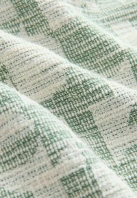 Tissu texturé avec un motif tissé de fils verts et crème, montrant des fils en relief et bouclés créant un motif en grille.