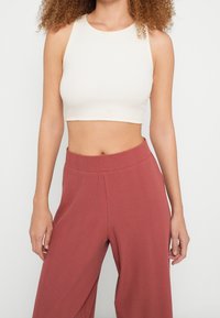 Femme portant un top court blanc sans manches et un pantalon ample taille haute couleur rouille sur un fond uni clair.