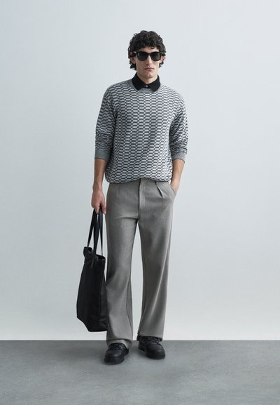 Suéter cinza com padrão e gola preta, calças plissadas cinza claro, sapatilhas pretas e uma mala tote preta. Texturas suaves por todo o conjunto.