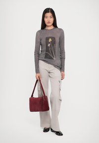 Femeie îmbrăcată cu un pulover gri cu un design floral, pantaloni cargo deschiși, balerini negri cu detalii metalice, ținând o geantă din suede bordeaux.