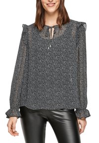 Blouse à motifs noir et blanc avec des manches longues, des épaules volantes et un col à nouer en tissu léger. Assortie à un pantalon noir brillant.