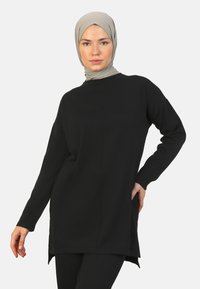 Zwarte langmouw tuniek met een ontspannen pasvorm, gemaakt van een zacht materiaal. Heeft zijsplitten en een ronde halslijn. Geaccentueerd met een lichtgekleurde hoofddoek.