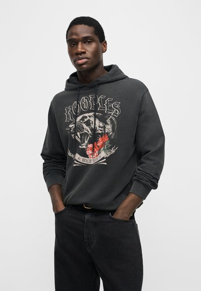 Homme portant un sweat à capuche noir avec une panthère rugissante, des fleurs rouges et le texte « RODEOLES WILD TOUR » imprimé sur le devant.