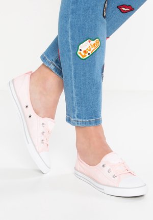 Ben iført blå jeans med farverige lapper og lyserøde lave lærredssneakers på hvid baggrund.