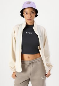 Kreem kergestusjakke, must lühike tee "ROXY" logoga, hallid spordipüksid "New Balance" tekstiga ja sireli värvi ämbri müts logoga.
