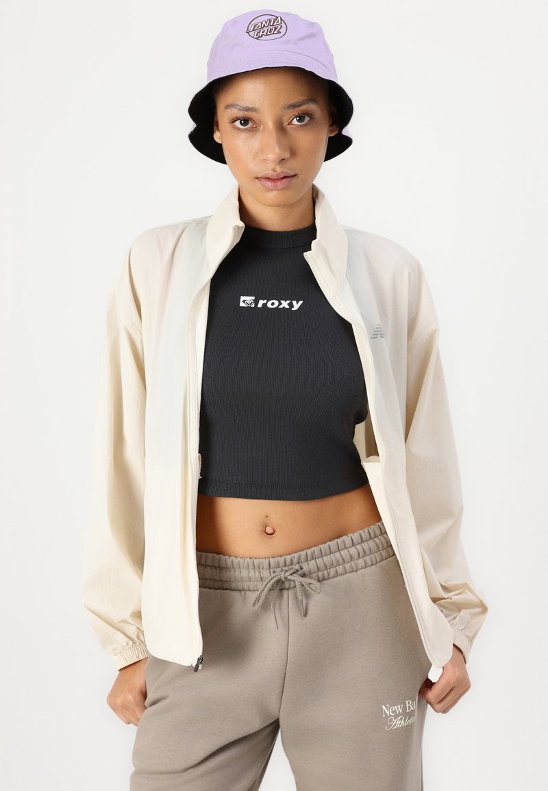 Veste légère crème, top noir court avec le logo "ROXY", pantalon de sport gris avec le texte "New Balance", et chapeau seau lilas avec logo.