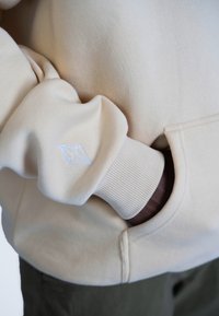 Felpa color beige con una texture morbida, polsini a coste, tasca frontale e un logo ricamato bianco sulla manica.