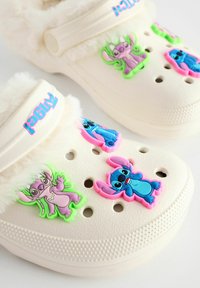 Sabots blancs avec doublure moelleuse, ornés de personnages en caoutchouc colorés et de trous de ventilation. Les accents incluent une bande de logo rose.