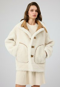 Witte fleece jas met bruin leren accenten, voorzien van een kraag, grote zakken en houten knopen. Zachte textuur, oversized pasvorm.