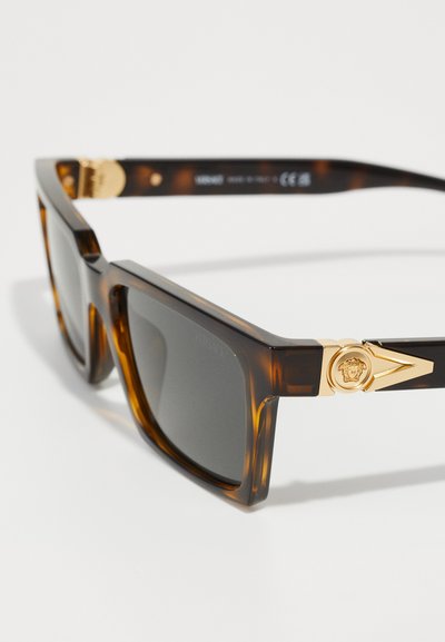 Tortoiseshell sunglasses with gray lenses, featuring gold hardware accents and a prominent emblem on the temple. Square-frame design.
