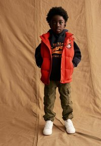 Gilet rembourré orange sur un hoodie marine avec du texte orange, pantalon cargo olive et baskets blanches. Fond en tissu beige.