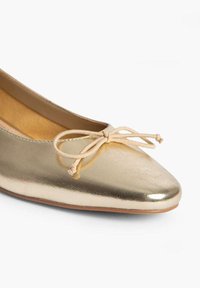 Ballerine en métal doré avec un bout pointu, ornée d'un nœud beige décoratif à l'avant et d'une surface lisse et brillante.