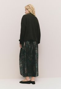 Schwarzer oversized Pullover, plissierter Blumenrock in dunklen Grüntönen und lila Nuancen sowie schwarze Ballerinas. Von hinten vor einem hellen Hintergrund zu sehen.