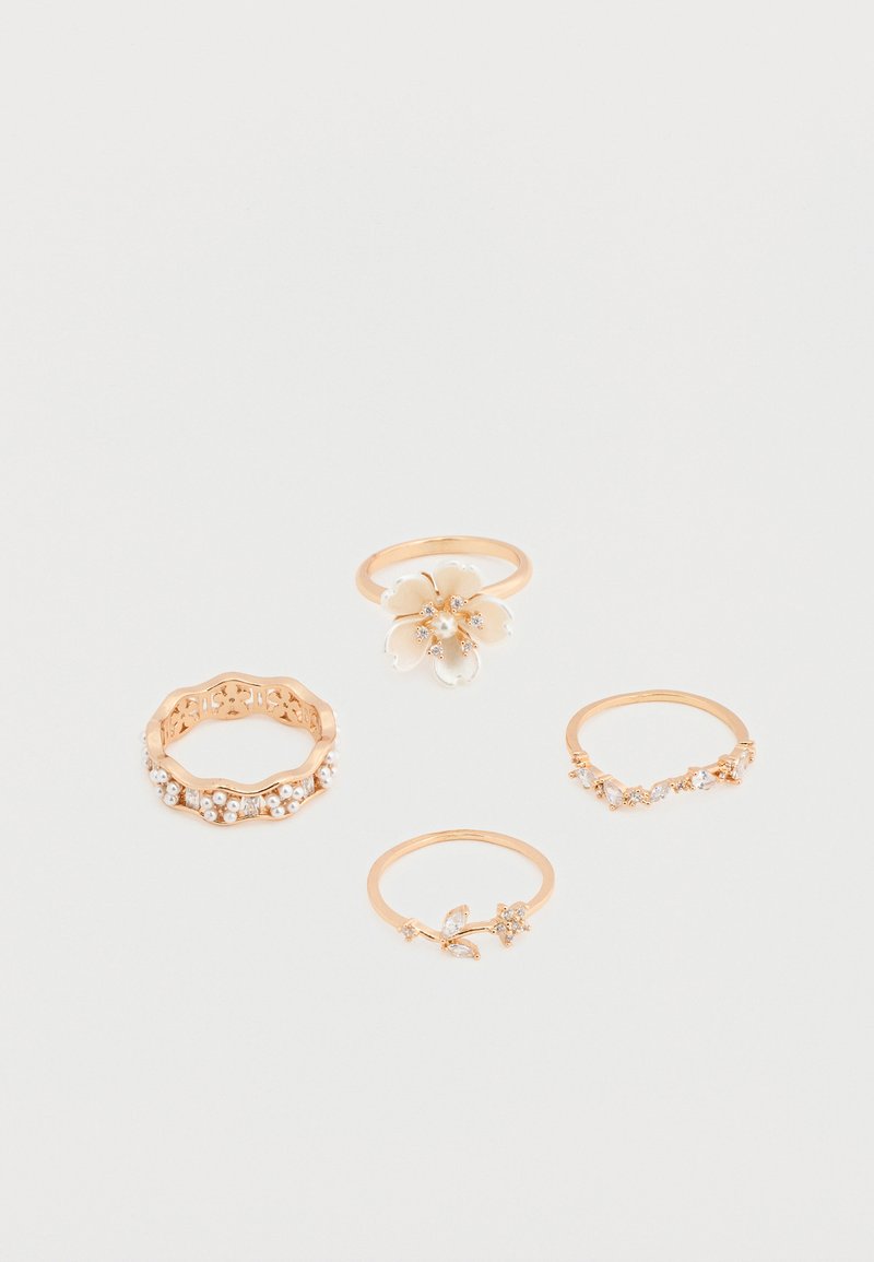 Cuatro anillos de oro con detalles florales y perlas dispuestos sobre una superficie blanca, destacando diseños delicados y acentos brillantes.