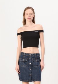Cropped top nero scollato con logo Calvin Klein, abbinato a una gonna di jeans con chiusura a bottoni frontali e due tasche anteriori.