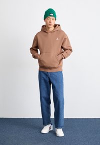 Sweatshirt à capuche marron avec une poche kangourou et un logo blanc, associé à un jean bleu et des baskets blanches avec des accents rouges et bleus.