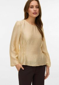 Blusa color crema stropicciata con maniche lunghe, scollo rotondo e dettaglio arricciato in vita. Abbinata a pantaloni neri. Tessuto strutturato.