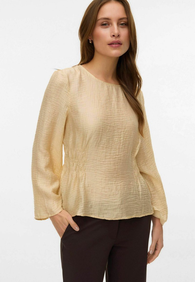 Blusa color crema stropicciata con maniche lunghe, scollo rotondo e dettaglio arricciato in vita. Abbinata a pantaloni neri. Tessuto strutturato.