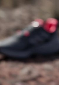 Scarpa sportiva nera con dettagli rossi, materiale testurizzato, design a profilo basso e sistema di lacci sicuro, adatta per l'uso all'aperto.