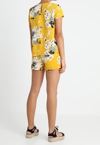 Gelbes Oberteil mit Blumenmuster und kurzen Ärmeln sowie passenden Shorts. Hat weiße, blaue und schwarze Blumenmuster. Getragen mit schwarzen Espadrilles.