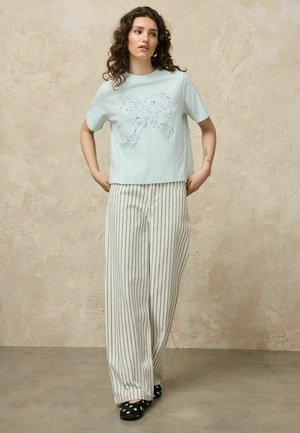 Femeie cu păr creț purtând o cămașă bleu deschis cu design cu fundă, pantaloni albi cu dungi largi și pantofi negri cu buline, stând cu mâinile în șolduri.