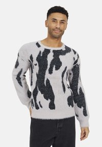 Pull en laine léger et fluffy de couleur gris clair avec un imprimé vache noir. Il possède un col rond, des manches longues et une texture douce. Coupe décontractée, associé à un pantalon sombre.