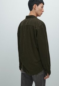 Camicetta in velluto a coste verde scuro con colletto, maniche lunghe e polsini con bottoni. Tessuto liscio, vestibilità rilassata e dettaglio con piega sul retro.