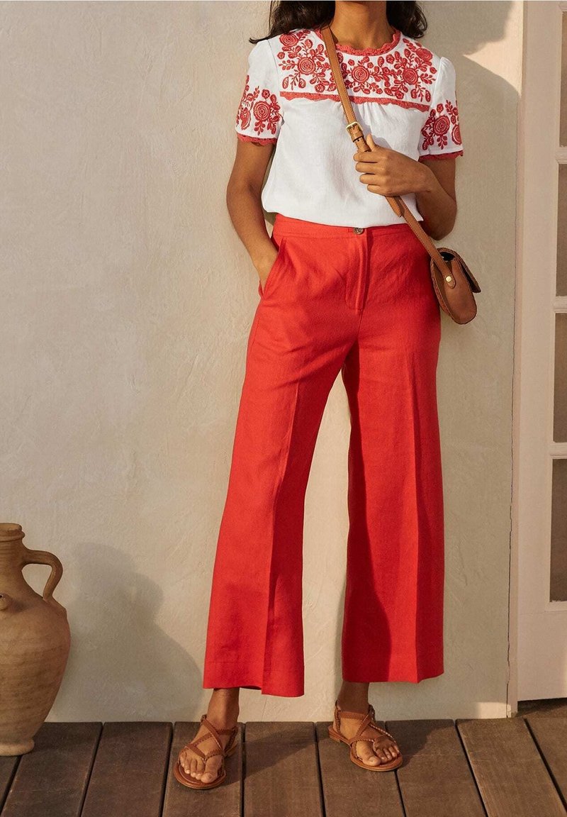 Femme portant un haut blanc avec une broderie florale rouge, un pantalon rouge taille haute à jambes larges, des sandales marron et un sac bandoulière marron.