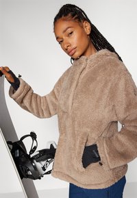 Brauner, flauschiger Fleece-Pullover mit hohem Kragen, Fronttasche und elastischen Bündchen. Getragen mit blauen Hosen, während eine Snowboard gehalten wird.