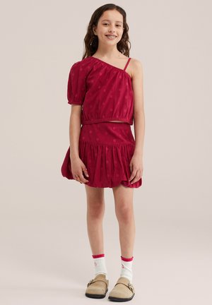 Egy lány áll és mosolyog, piros pöttyös egyrészes felsőt, hozzá illő szoknyát, fehér zoknit és bézs slip-on cipőt visel.