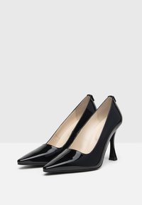 Tacones altos de charol negro con puntera puntiaguda y un tacón delgado y en ángulo. El interior cuenta con forro beige. Diseño clásico y elegante.