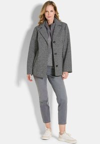 Femme aux cheveux blonds portant une tenue grise superposée comprenant un manteau texturé, un pantalon court et des baskets blanches, se tenant devant un fond uni.