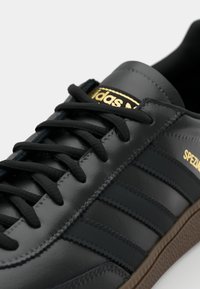 Sneaker in pelle nera con tre strisce scure, logo "Adidas" oro, lacci neri piatti e suola in gomma marrone. Design classico con dettagli testurizzati.