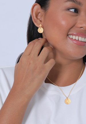 Goldene Ohrringe mit runden, strukturierten Anhängern und einer passenden goldenen Halskette mit einem ähnlichen Anhänger, getragen zu einem weißen Shirt.