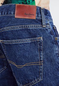 Jeansi iz denima v temno modri barvi, z oranžnimi šivi in rjavim usnjenim logotipom z napisom "Pepe Jeans London" na pasu.