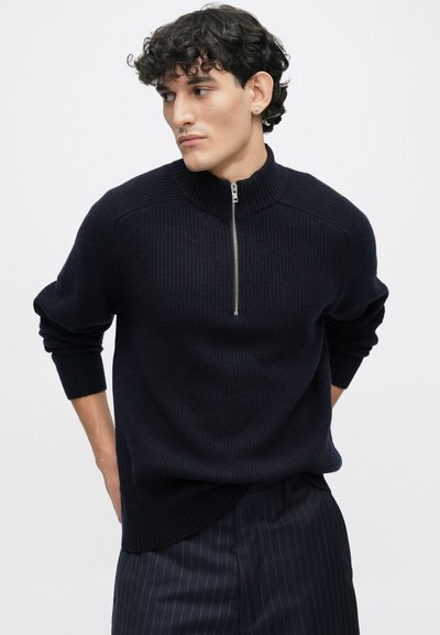 Dark navy knit sweater with a high collar and half zipper, featuring ribbed texture and long sleeves. Paired with pinstriped trousers.