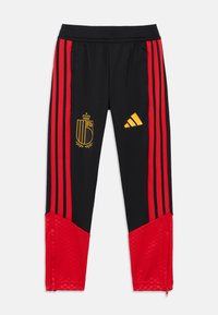 Calças atléticas pretas com riscas vermelhas nas laterais, apresentando um emblema dourado e o logótipo da Adidas. A textura é lisa com detalhe de fecho perto dos tornozelos.