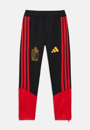 Schwarze Sporthose mit roten Seitenstreifen, einem goldenen Emblem und einem Adidas-Logo. Die Oberfläche ist glatt, mit Reißverschlussdetails in der Nähe der Knöchel.