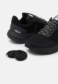 Baskets noires en mesh et en caoutchouc, avec une surface texturée, des accents blancs et deux paires de lacets noirs à côté.