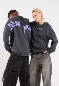 Two models wear grey hoodies with "New York" in purple on the back and front. They pair with distressed dark jeans and textured fabric.