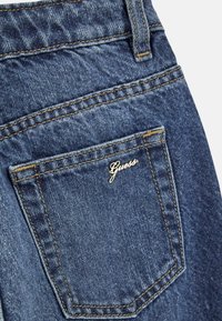 Poche arrière d'un jean en denim avec un logo doré "Guess". Tissu bleu foncé, couture visible et finition texturée.