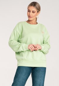 Jasnozielona bluza z luźnym krojem i ściągaczami. Posiada mały, graficzny wzór napoju na lewej piersi. Miękka faktura.