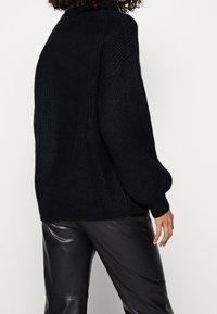 Persoon in een zwarte dikke gebreide sweater en zwarte leren broek, met de rug naar de camera gekeerd en krullend haar, tegen een eenvoudige witte achtergrond.