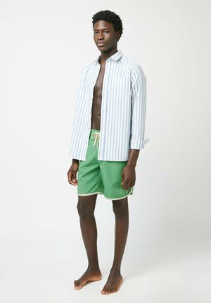 Camicia a righe blu e bianche con bottoni, pantaloni da bagno verdi con bordi bianchi, modello con capelli corti, a piedi nudi, in piedi su uno sfondo neutro.