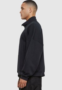 Črna ženska sweatshirt jakna z zadrgo in visokim ovratnikom, ohlapnega kroja ter dolgimi rokavi. Ima sprednji žep in rebraste manšete na robu.