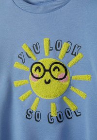 Világoskék pulóver, jól látható, plüss sárga nap mintázattal, napszemüveggel, rózsaszín pofival és fekete felirattal: "OLYAN JÓL NÉZEL KI!"