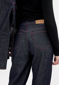 Temno modri džins hlače z rahlo ohlapnim krojem, z rdečim šivom, usnjeno oznako na pasu in klasičnimi zadnjimi žepe.