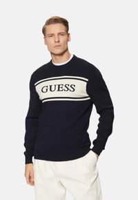 Suéter azul marino con franjas crema y el logo "GUESS" en el pecho. Puños y dobladillo acanalados, cuello redondo, fabricado en un suave tejido de punto.